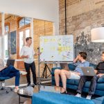 Three men sitting while another man presents in front of a whiteboard